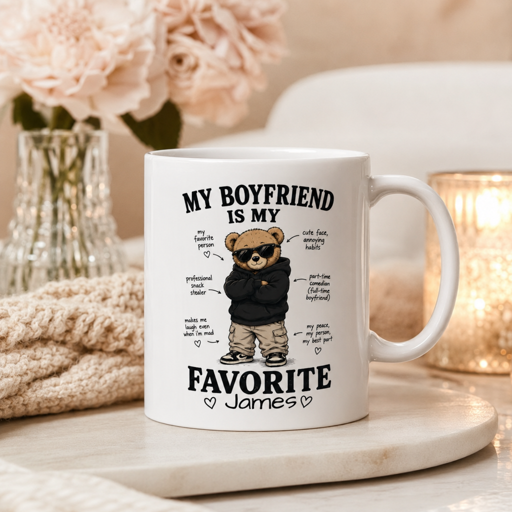 A white ceramic mug with a custom cartoon bear graphic and the text 'MY BOYFRIEND IS MY FAVORITE James' styled on a marble tray with a knitted blanket and soft background decor.