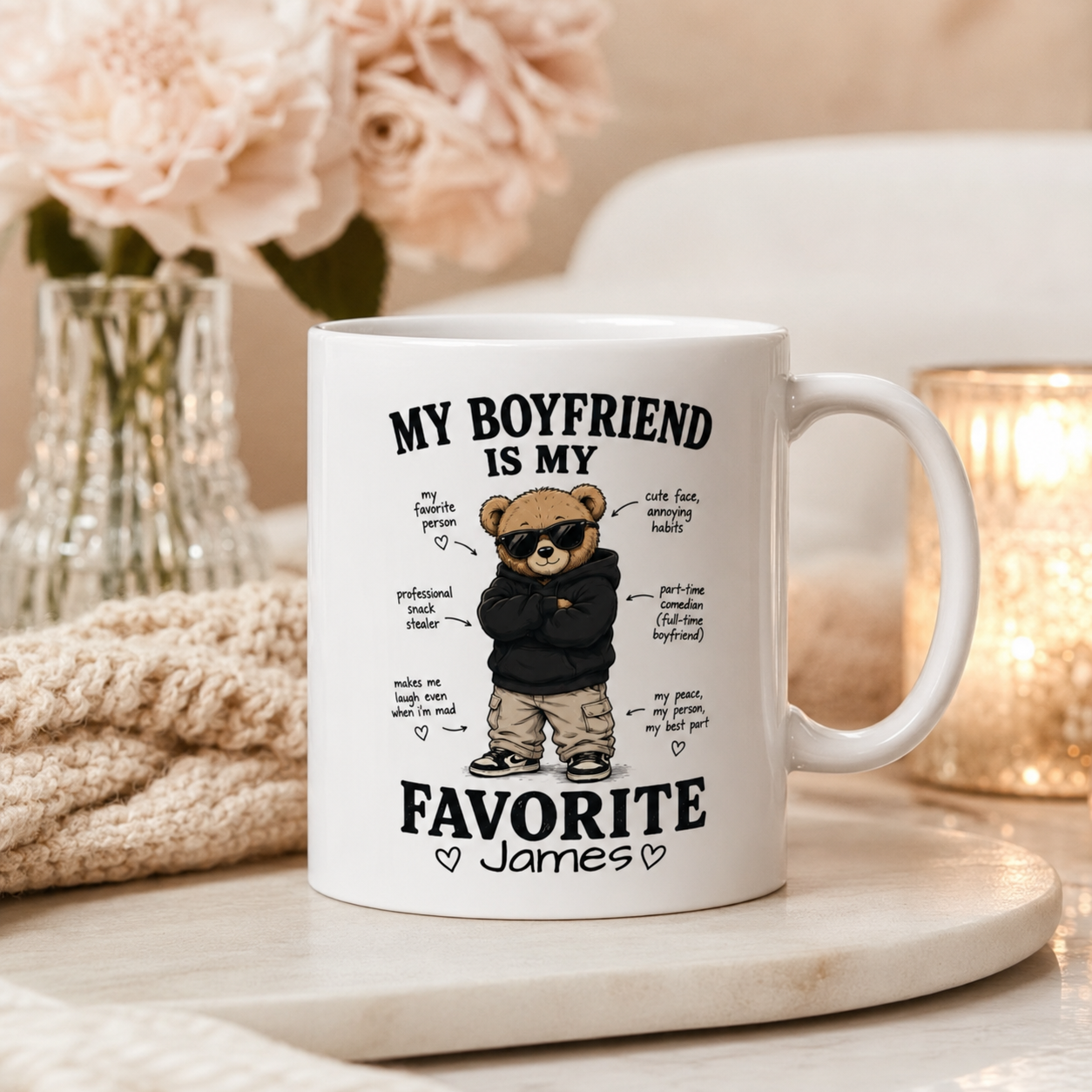 A white ceramic mug with a custom cartoon bear graphic and the text 'MY BOYFRIEND IS MY FAVORITE James' styled on a marble tray with a knitted blanket and soft background decor.